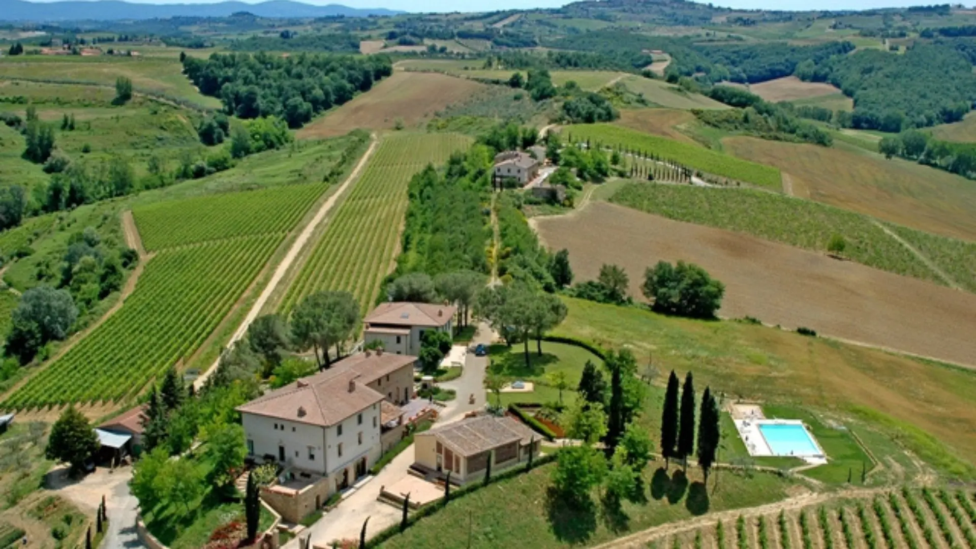 In the San Gimignano region, in Tuscany, you can see the Poderi Arcangelo winery and vineyards from above. The vineyards are aplenty and the surrounding lands are green.