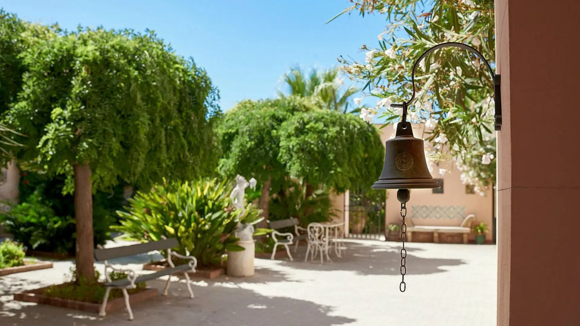 The courtyard of the Baglio Ingardia winery. You can see a few benches, trees, as well as the walls of the estate. There is also a regular old metal bell with a clapper and chain on the wall of the winery.