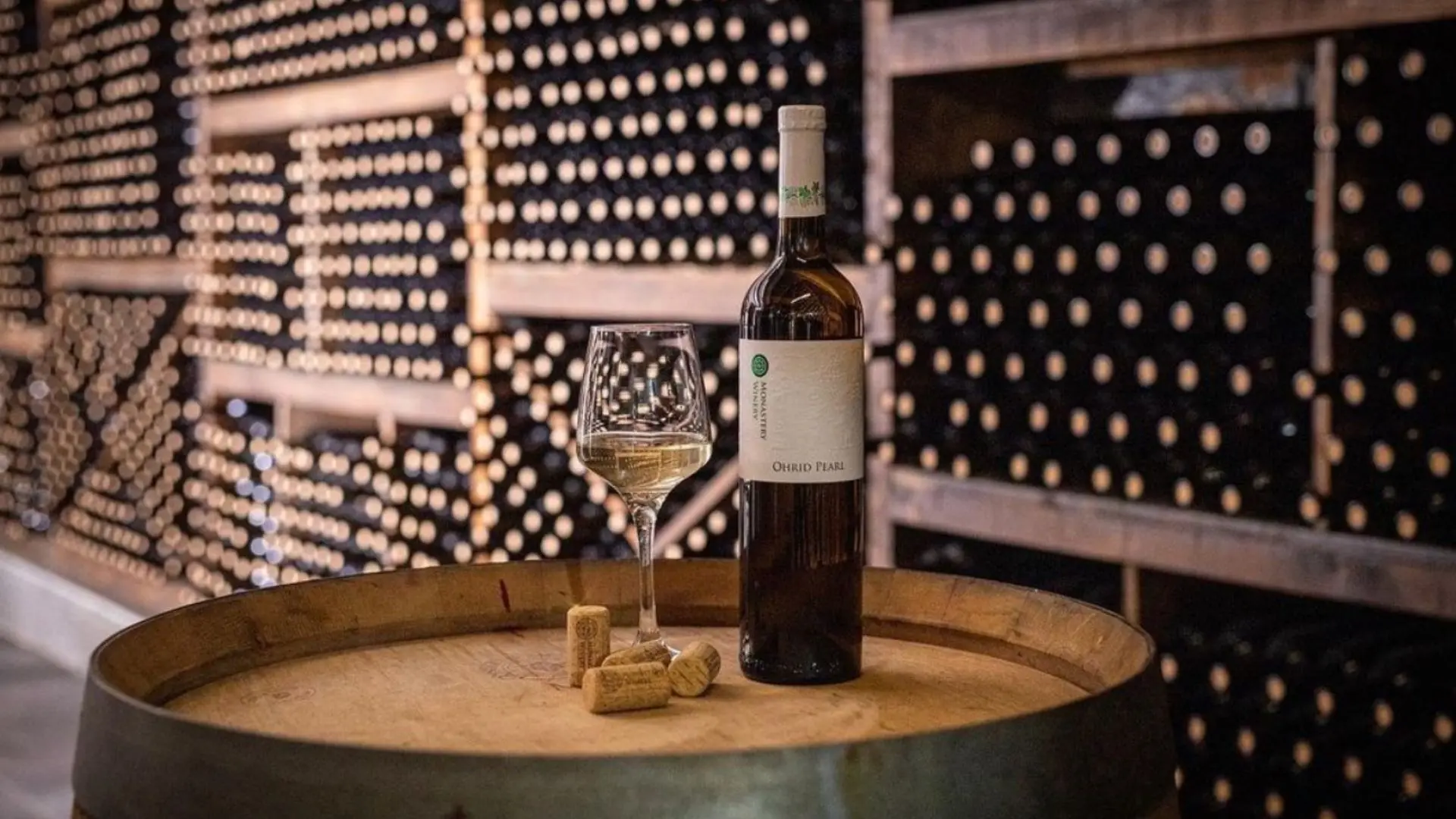 A large number of wine bottles on a shelf at the Monastery winery. There is a bottle of white wine on a barrel. A glass of white wine can also be seen, and four corks.