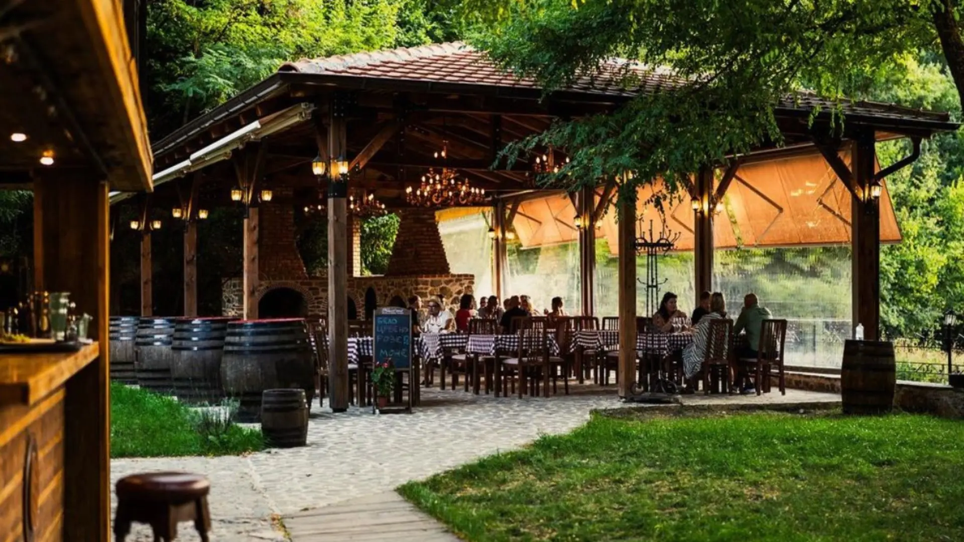 A typical wine tasting experience at the Monastery winery. There is a patio with a tiled roof above. People can be seen, seated at a table. There is a stone grill behind them and wine barrels to the left of them.