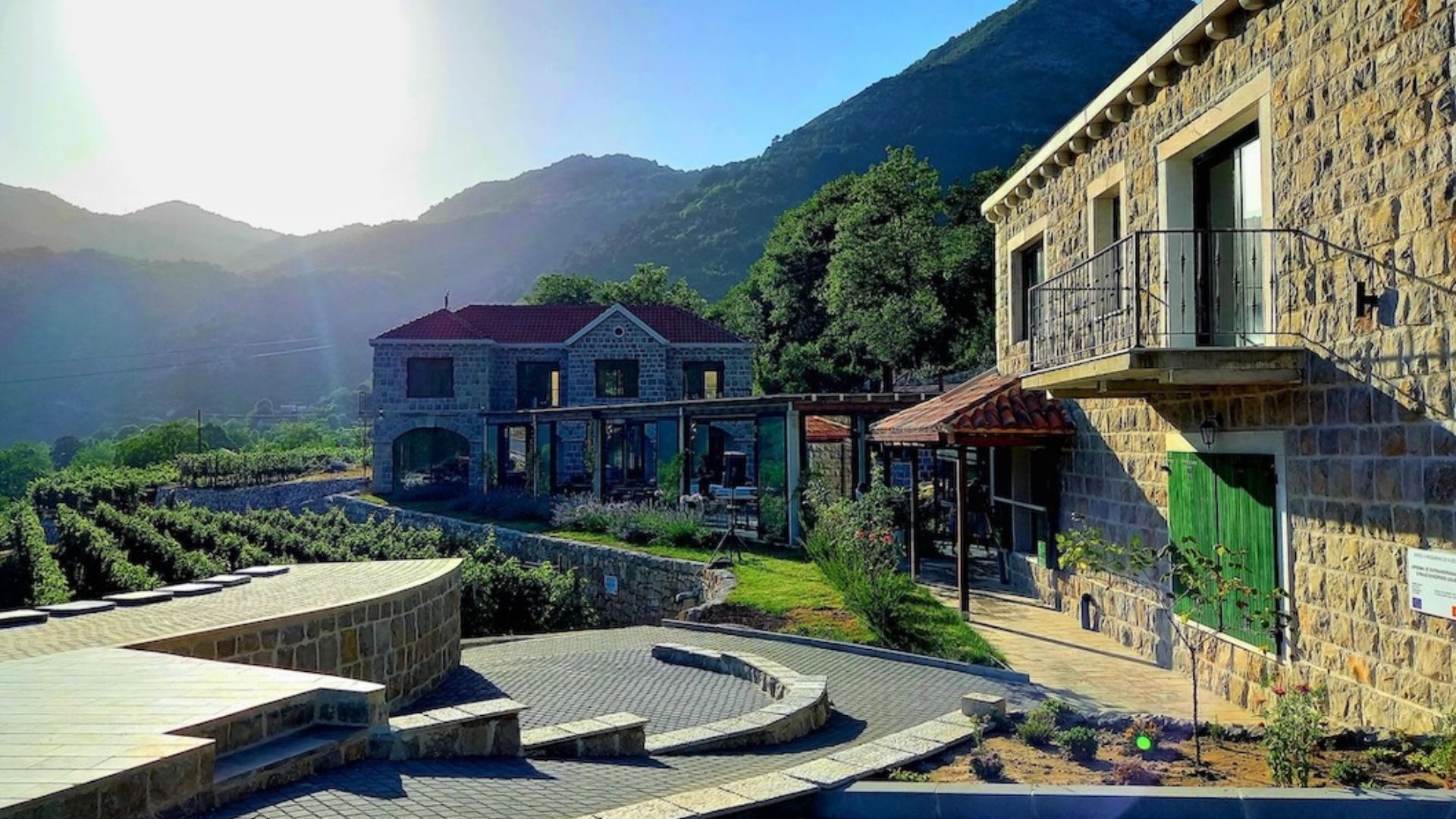 The image shows two old houses with vineyards to the left. To the right, one can see trees. The background is full of hills and a blue skyline.
