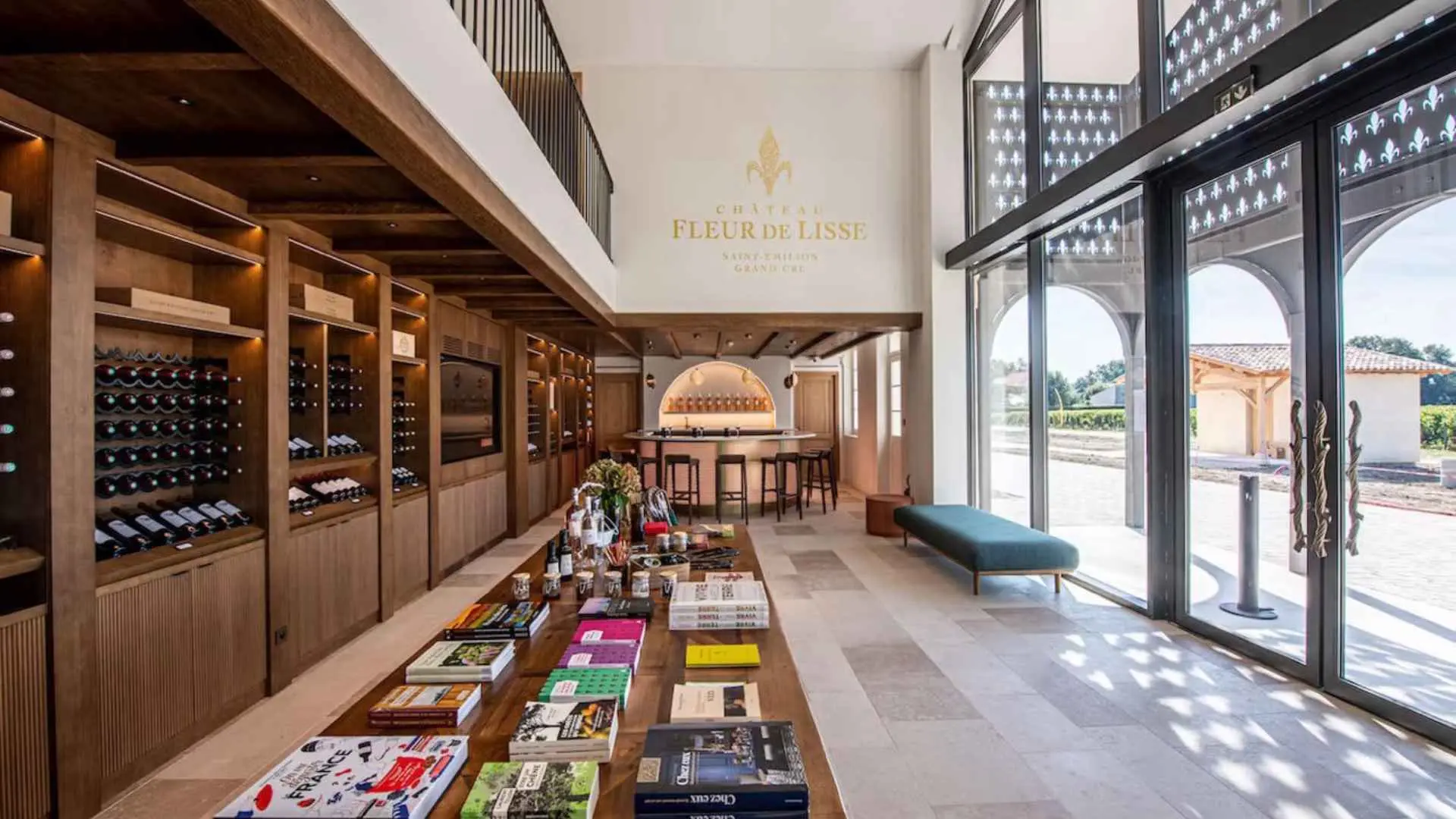 Château Fleur De Lisse winery interior. There is a table full of books and magazines. There are wine bottles to the left, and a bar in the back. The right part of the image has a large glass door and window.