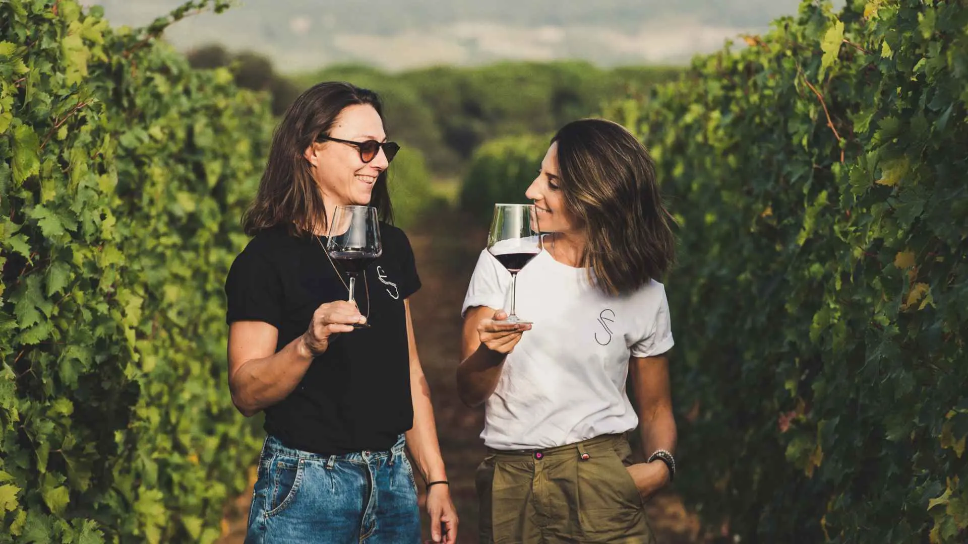 The image shows the two sisters, owners of the Le Vigne di Silvia winery, having a glass of red wine in their vineyard, during a late afternoon.