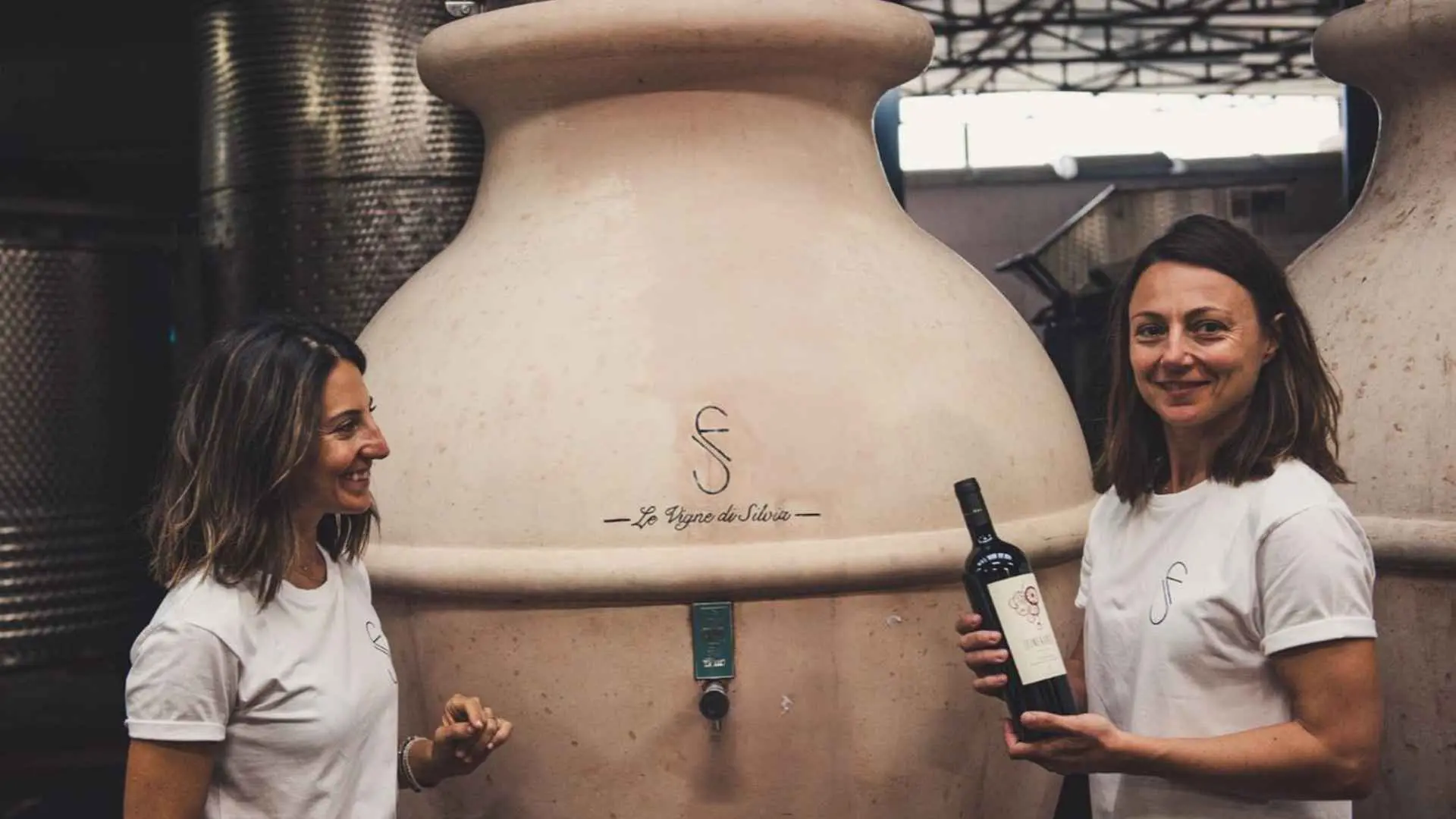 The image shows the two owners of the Le Vigne di Silvia winery. They are positioned next to a huge wine tank.