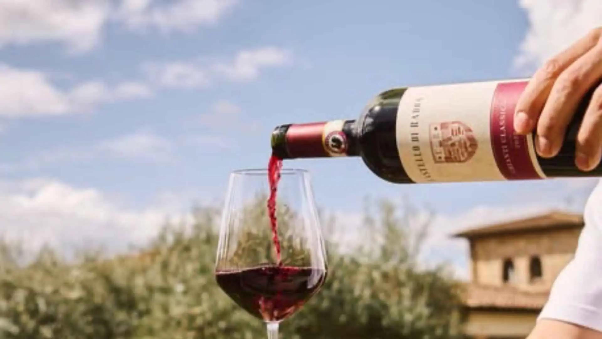 The image shows a person pouring red wine into a glass at the Castello di Radda winery. The sky behind them is a bit cloudy.
