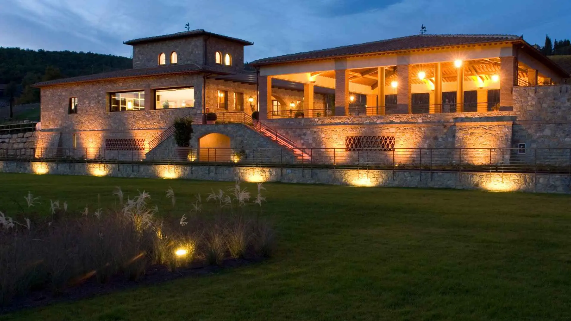 The image shows the Castello di Radda winery and courtyard right after a sunset. The lights are warm and the lawn cleanly cut.