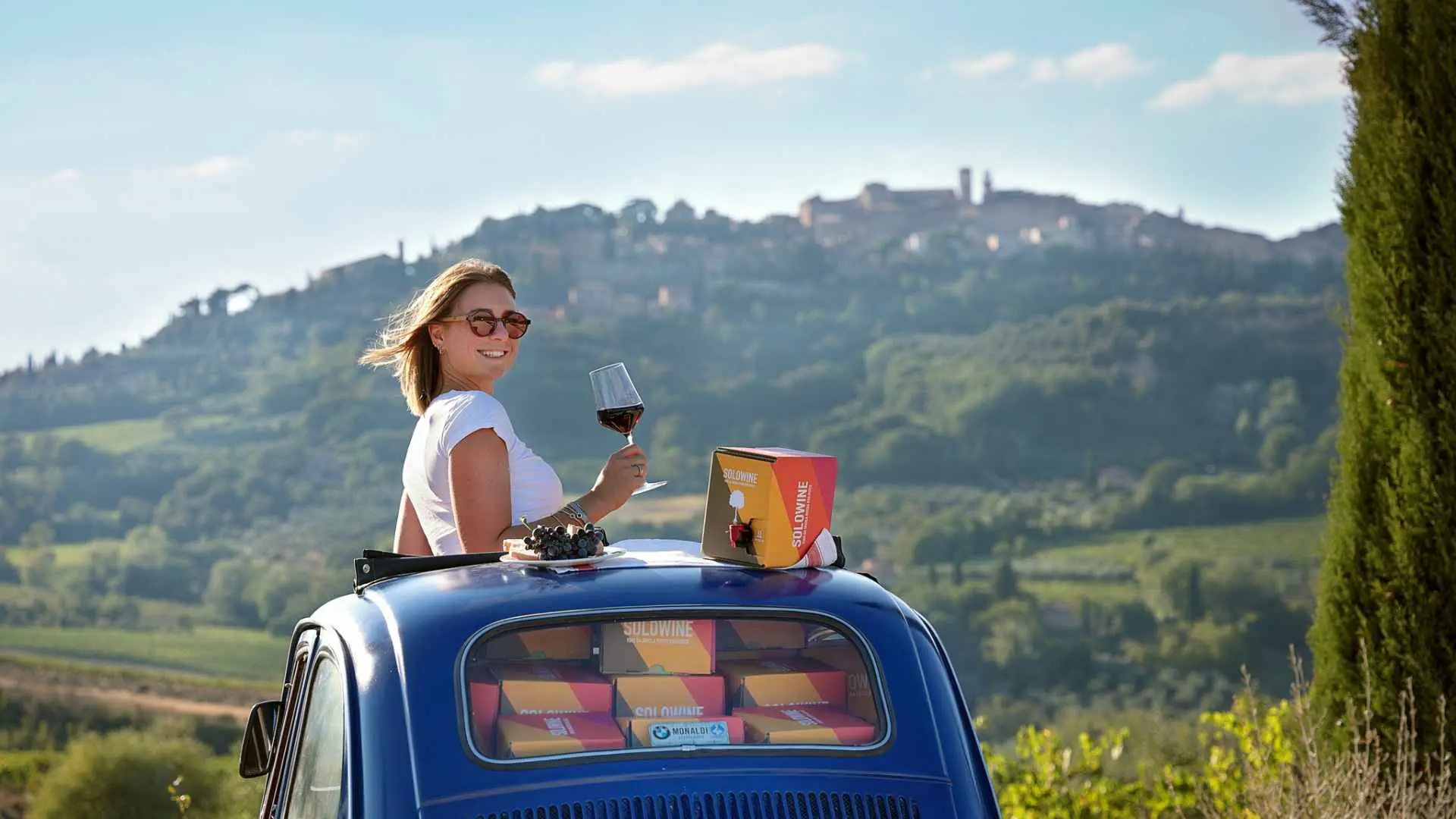 An image showing a woman in a convertible Fiat 600. The woman is holding a glass full of red wine. In the background, you can see the Salcheto winery in the Montepulciani region in Italy.