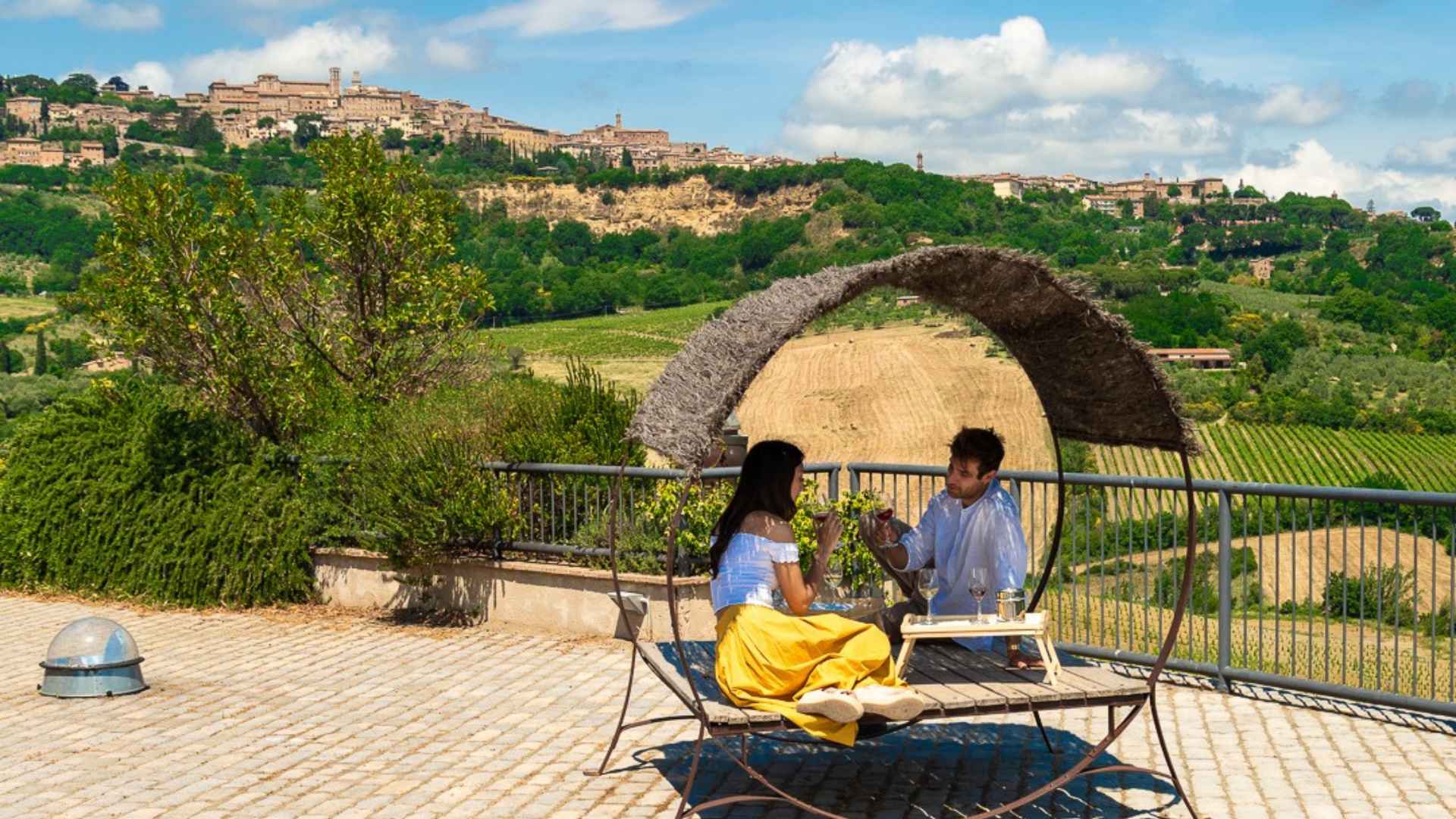 The image shows a man and woman having wine under a cover of shade. Behind them is the Salcheto winery vineyard.