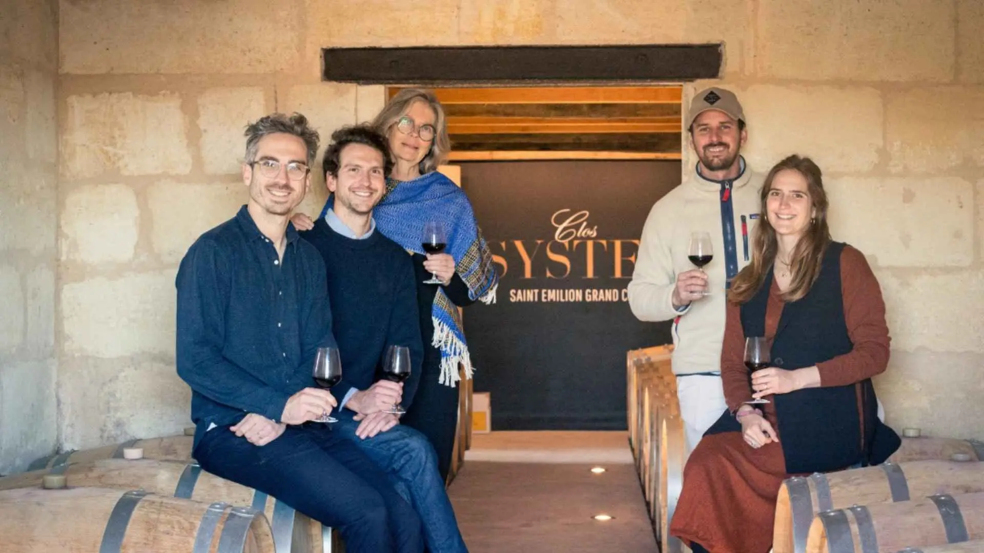 The image shows Clos Systey workers seated on top of wine barrels in a stone room. Behind them is a sign with the brand name.