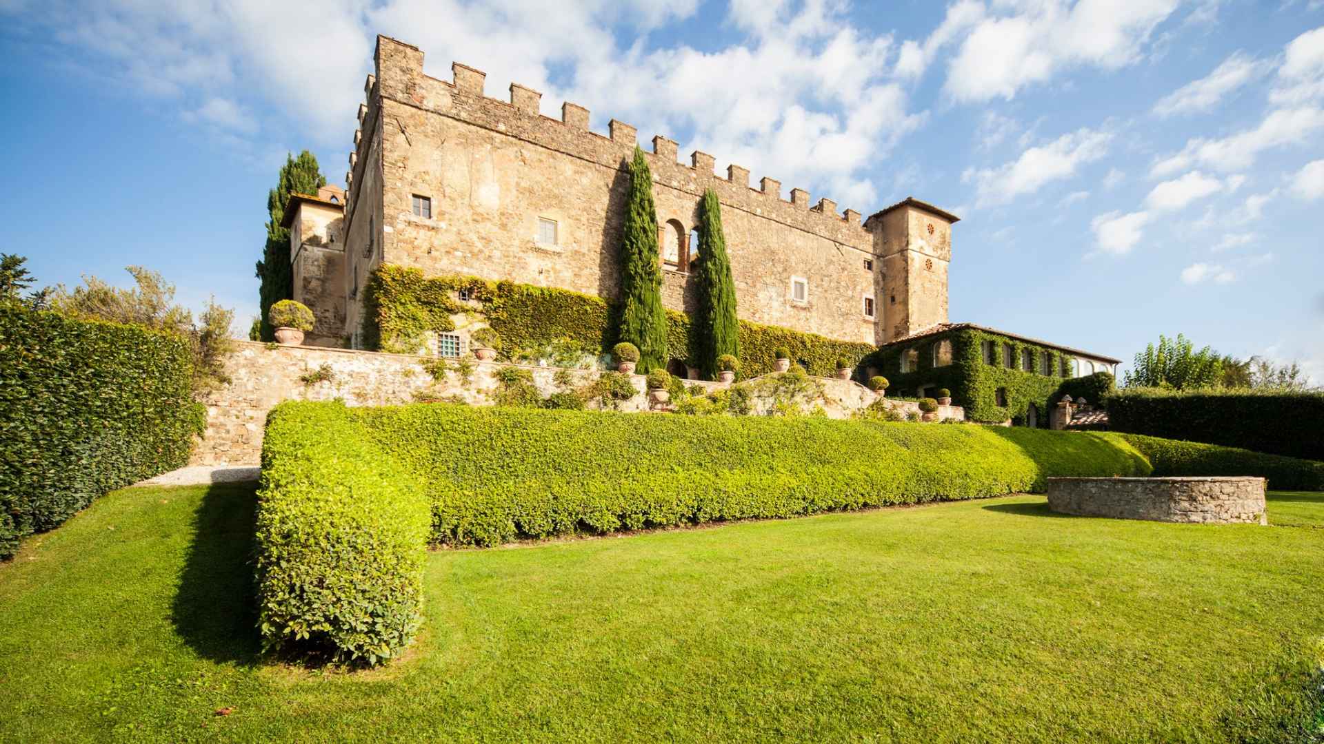 The image shows the castle and winery of Castello della Paneretta. There is an overcast sky above and beautiful green shrubs and grass in front.