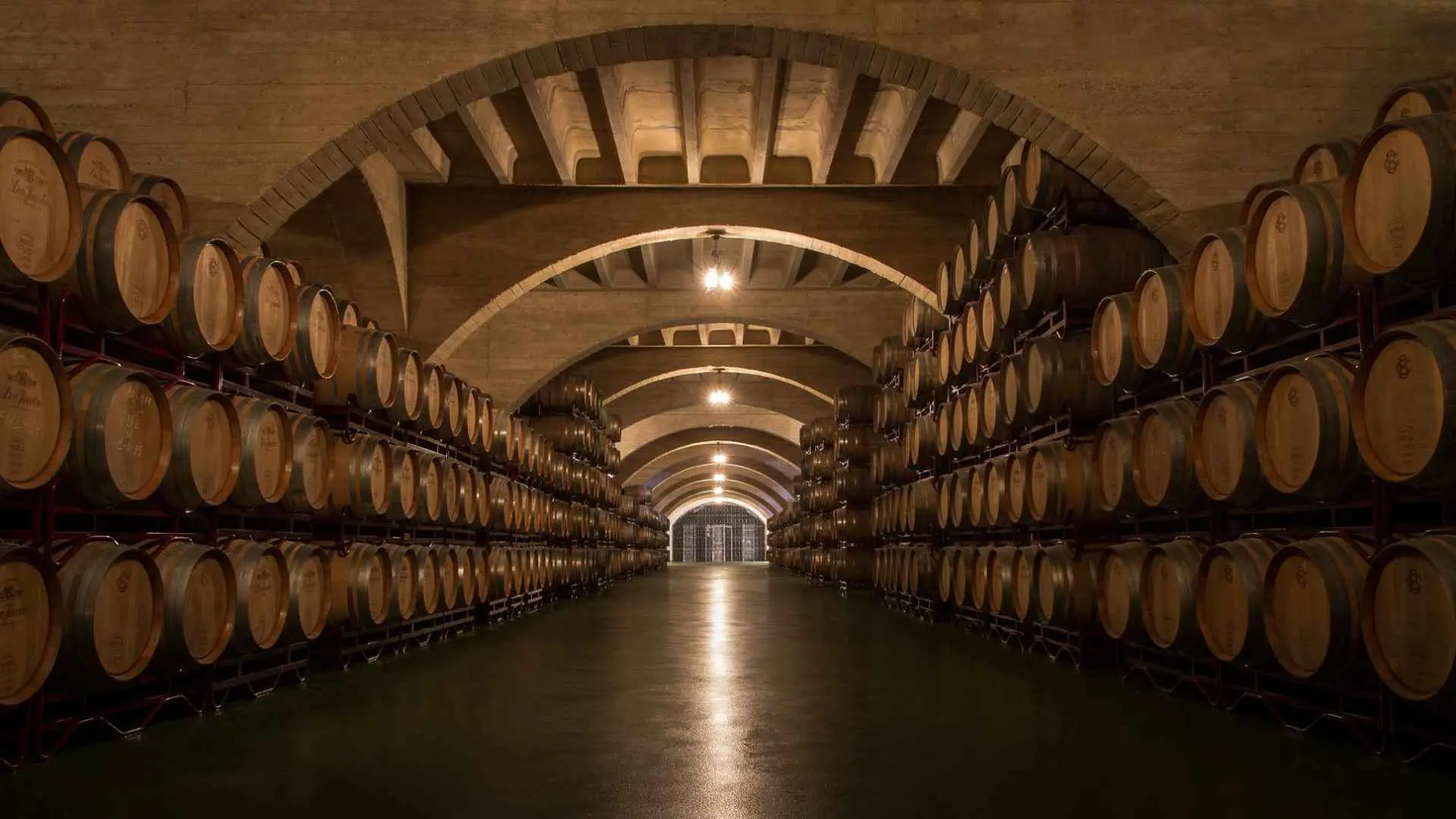 The image shows the wine cellar of the Bodegas Corral. It has barrels on both sides, stacked sideways one atop another. The cellar has arches and is lit with a warm light.