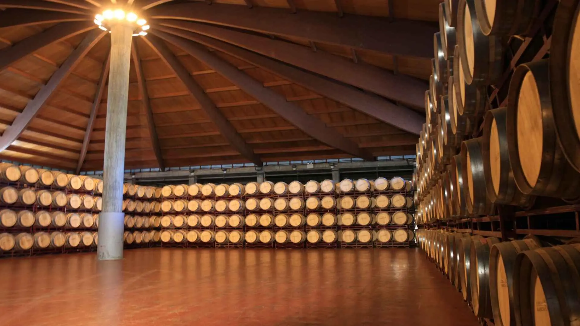 The image shows a wine cellar of the Bodega Jaume Serra. There are four columns of wine barrels stretching on each wall of the room. There is a wooden pillar in the middle, where the light is also hoisted.