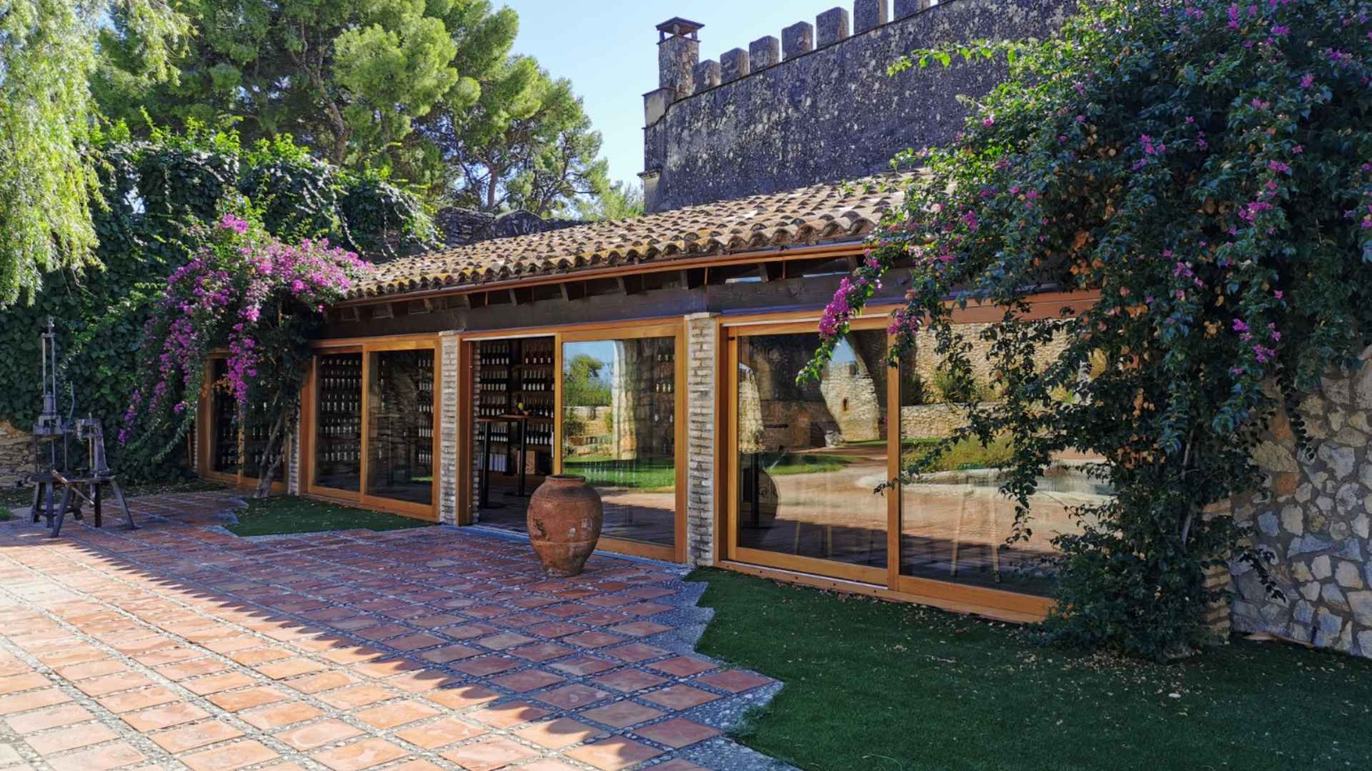The building of the Bodega Jaume Serra winery. It is a beautiful stone building with large glass windows. There are trees and flowers in the image, as well as a grass lawn and stone pathway.