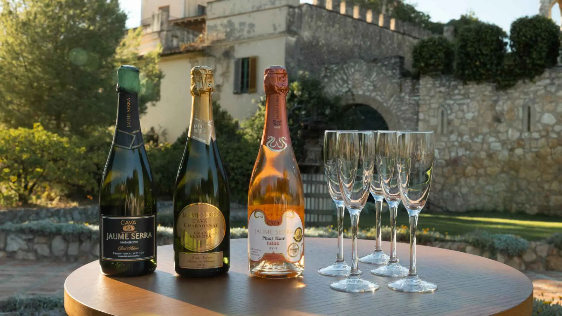 Three very distinct bottles of wine and four wine glasses, overlooking the stone courtyard of the Bodega Jaume Serra.