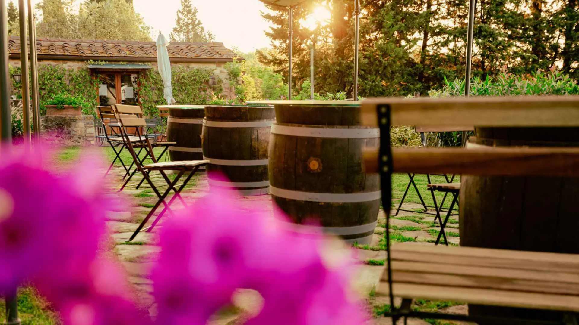 The image has wine barrels as tables and wooden chairs in a beautiful garden. The time of day is the afternoon and there is a purple flower in front of the image, out of focus.