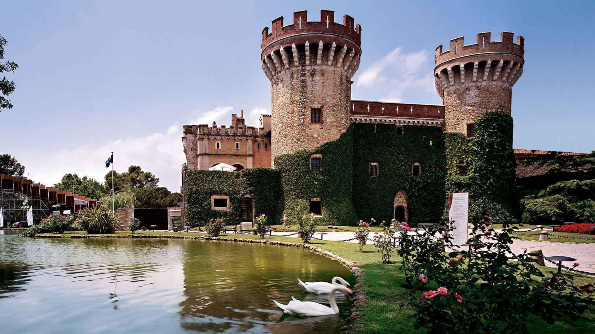 Nou Celler Perelada castle can be seen in the image. It is an old stone building, withe vines creeping up its main wall and two towers. A pond can be seen in the lower left part of the image. The sky is blue.