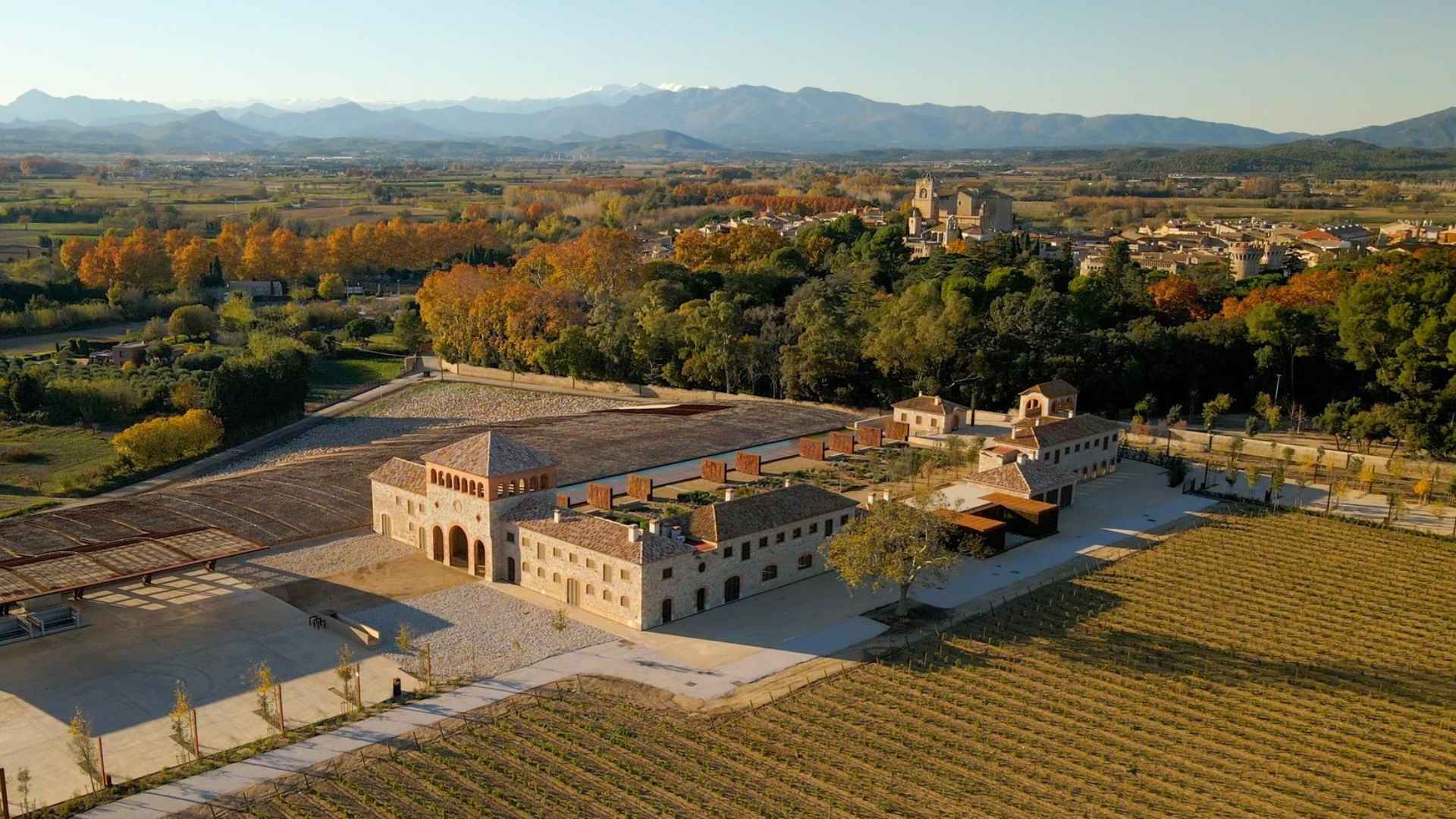 The Nou Celler Perelada winery and vineyards can be seen on the image. There is also a smallish forest with yellowing leaves. The time of year is autumn and the sun is low. The sky is blue.