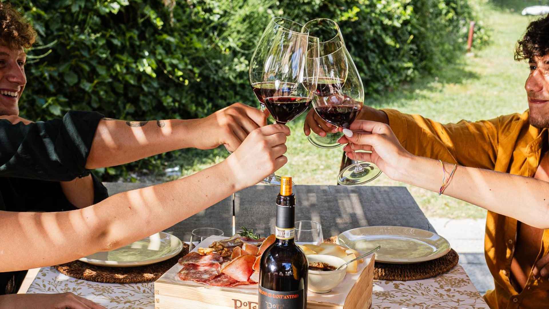 Molino di Santa Antimo wine tasting. The image shows four people and wine glasses. They are toasting. There is food on th etable and greenery can be seen in the background.