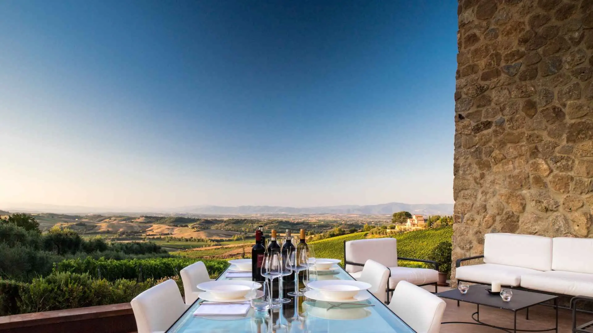 A set table with four different wine bottle at the Icario winery. An old stone building can be seen to the right, while the balcony overlooks the vineyards. The image was taken during a sunset.