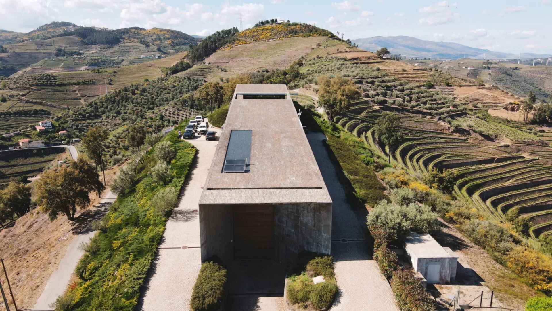 The Adega H.O winery building can be seen in the middle of the image. It is flanked by its vineyards, while there is an overcast sky in the background.