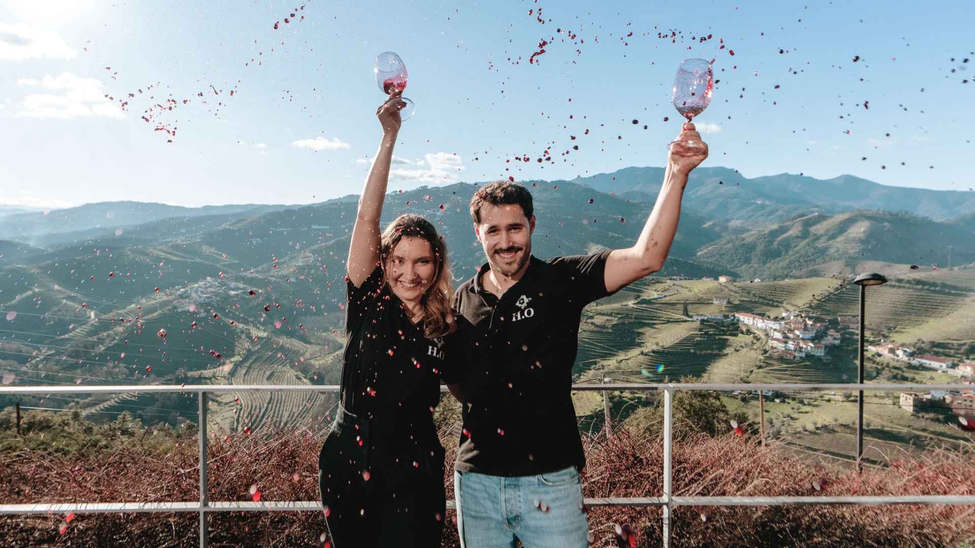 Two people can be seen, their arms held high, with half-empty glasses of wine. Red wine can also be seen everywhere on the image, as if they spilled it intentionally for the shot. 