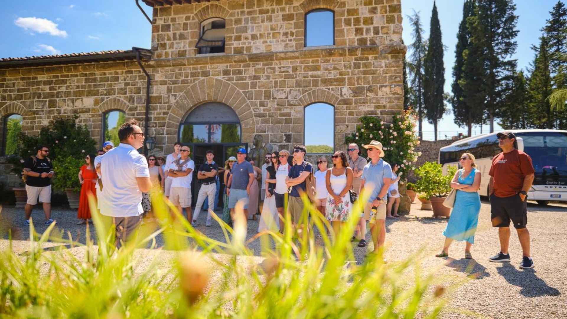 The image shows people in front of the Chianti Vineyards. The people are facing a guide. Behind them is the Chianti Vineyards building, an old structure, as well as a bus, trees, and a blue skyline.