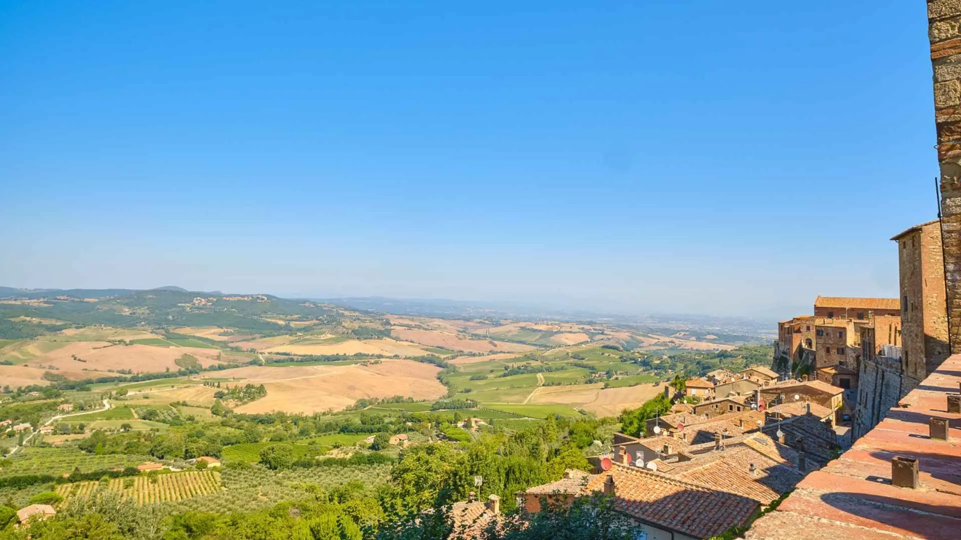 The view shown from the walls of the Montepulciano walls in Tuscany, Italy. It overlooks vineyards, various fields, as well as a blue skyline.