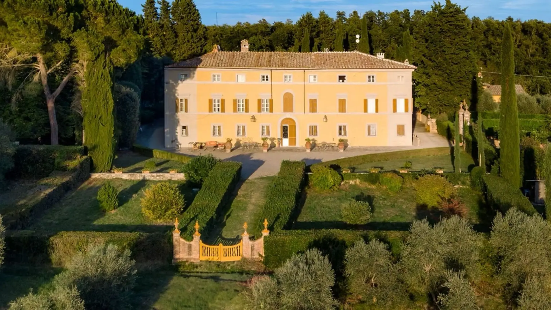 This old estate looks quite the part and the image shows its glory during the golden hour of the day. In front of the estate, one can see all sorts of trees, a garden, as well as a gate, in the color of the estate, which is yellow.