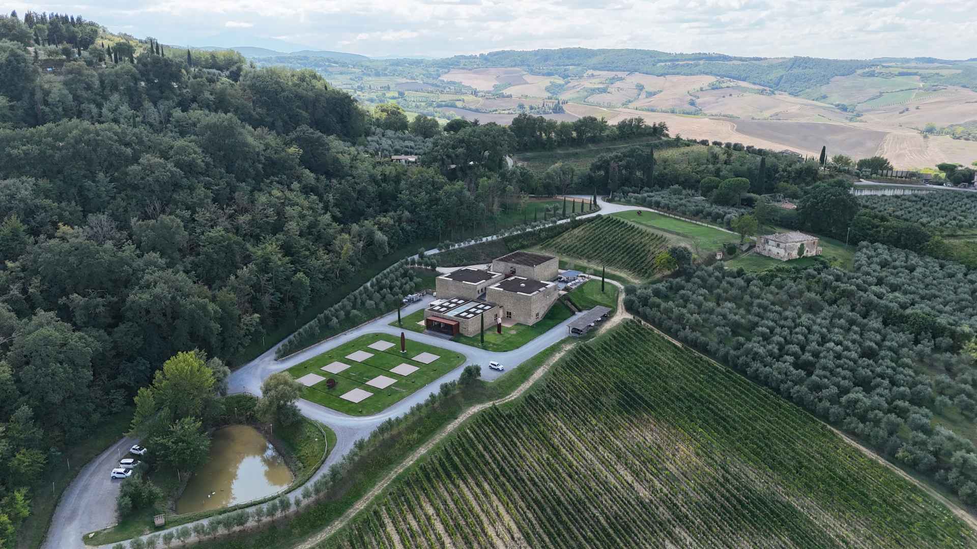 Icario is a modern winery in Tuscany, a region known for its wine-making. The image shows a vineyard in the foreground. The center of the image hosts the winery, while it is flanked by a forest to the left and more vineyards to the right. The background shows hills and an overcast skyline.