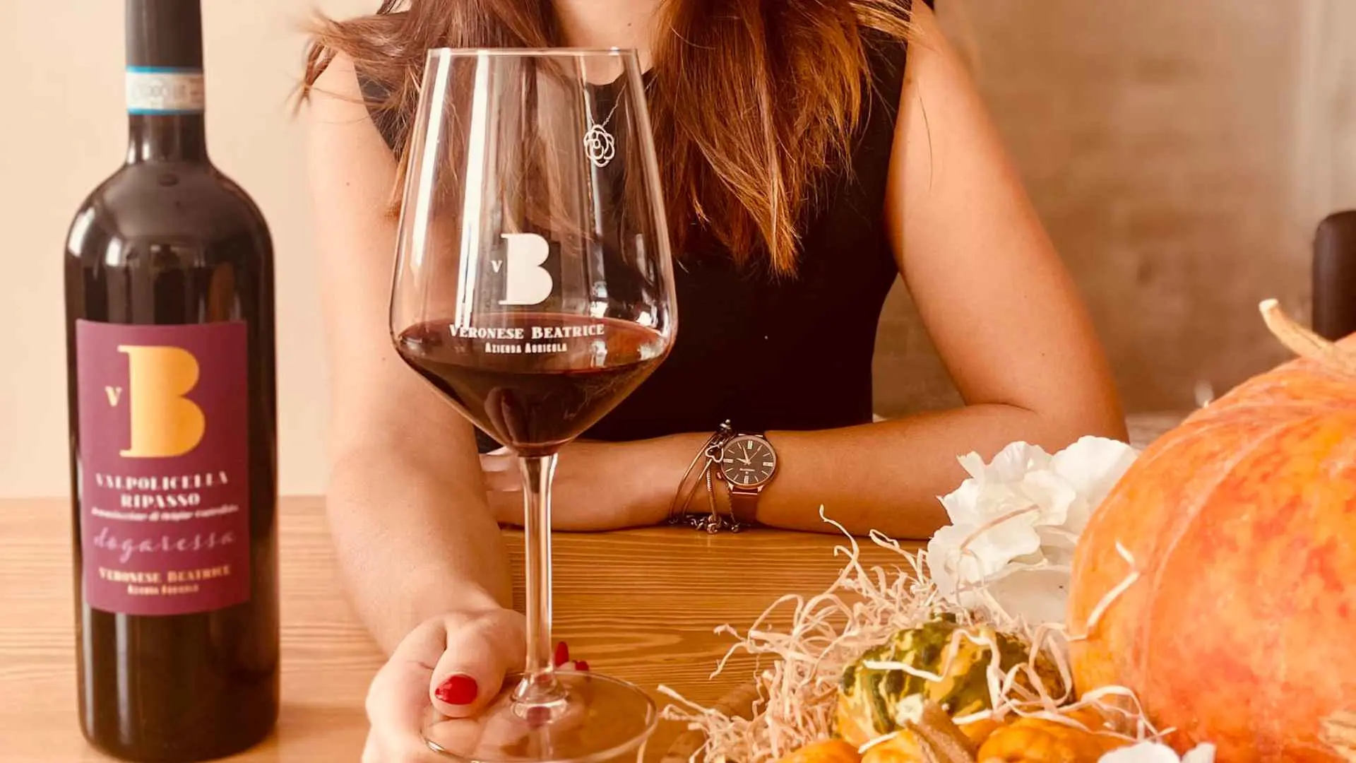 A woman holding a glass of red Veronese Beatrice wine. The bottle is to left and there is food to the right.