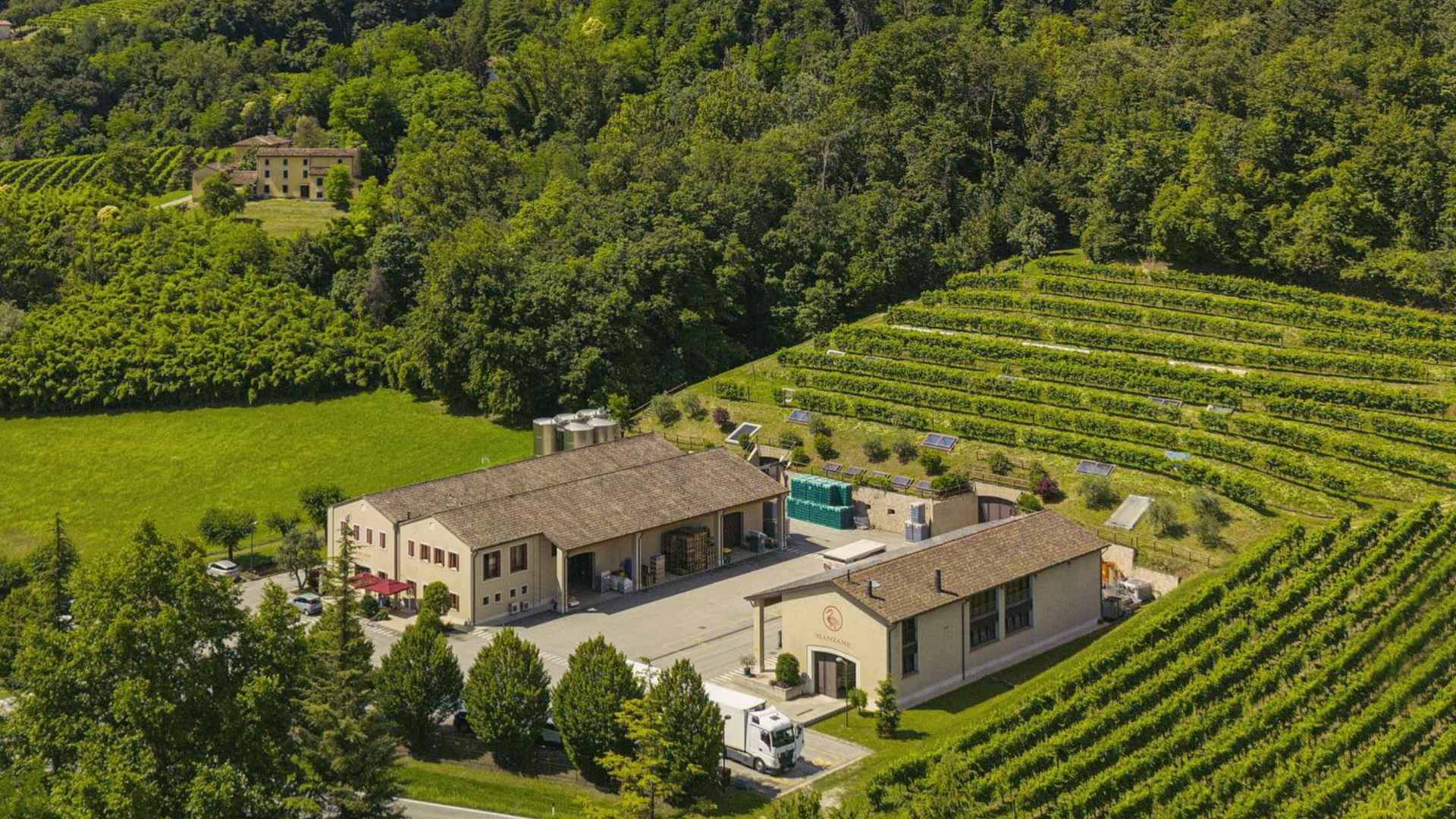 The image shows the La Manzane winery, from above. It is surrounded by vineyards on the right side, a forest in the back, while it has a beautiful green lawn to the left.
