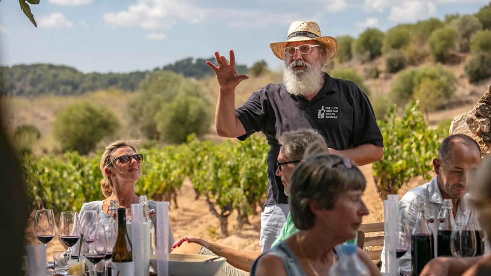 The image shows people having a wine tasting at the Bell Cros winery. They are having a wine tasting while a guide is explaining stuff to them. The background has vineyards and a blue sky.