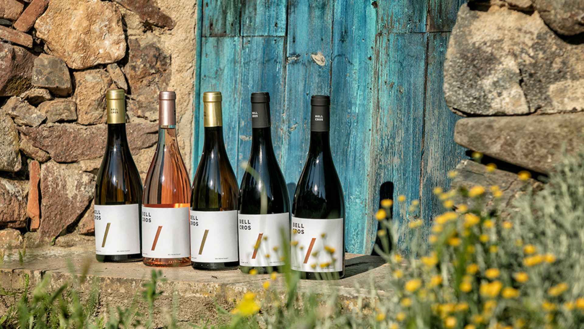 The image shows various Bell Cros wine bottles in front of an old, blue door, next to various stone walls, and a porch.